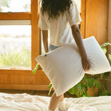 Person holding a Stitchco pillow in a bright room with a window and plants.