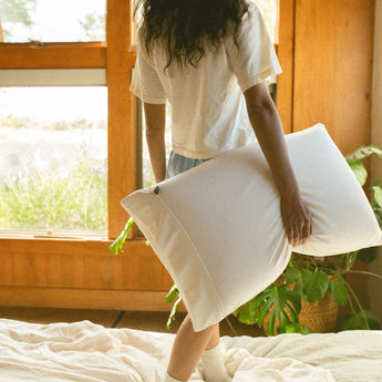 Person holding a Stitchco pillow in a bright room with a window and plants.