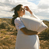 Woman in a white dress holding a Stitchco pillow in a desert landscape with mountains and blue sky.