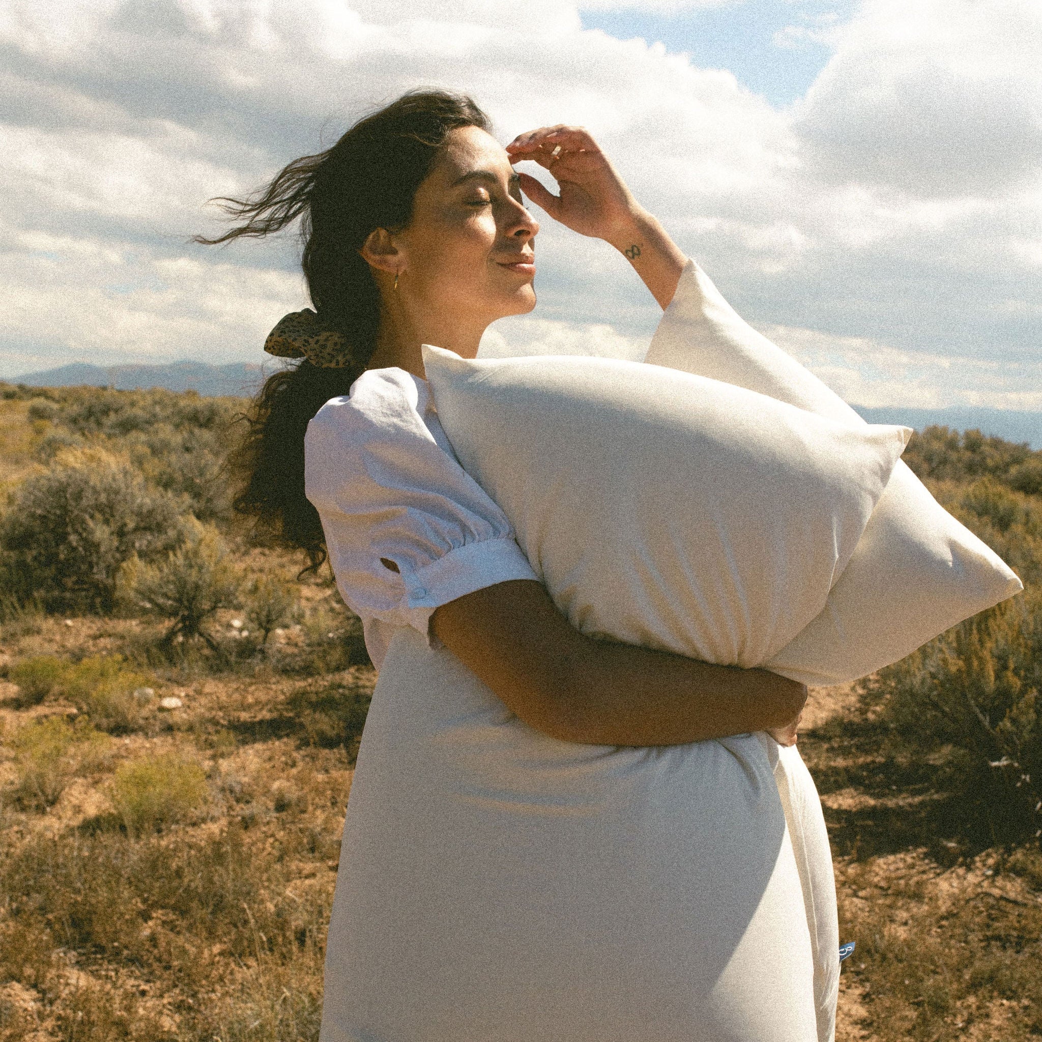 Woman in a white dress holding a Stitchco pillow in a desert landscape with mountains and blue sky.